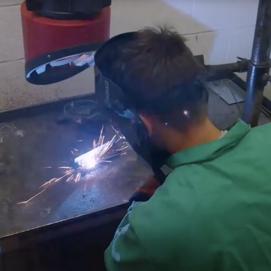 Man welding metal on a workbench sparks flying wearing protective green jacket and welding helmet indoors.