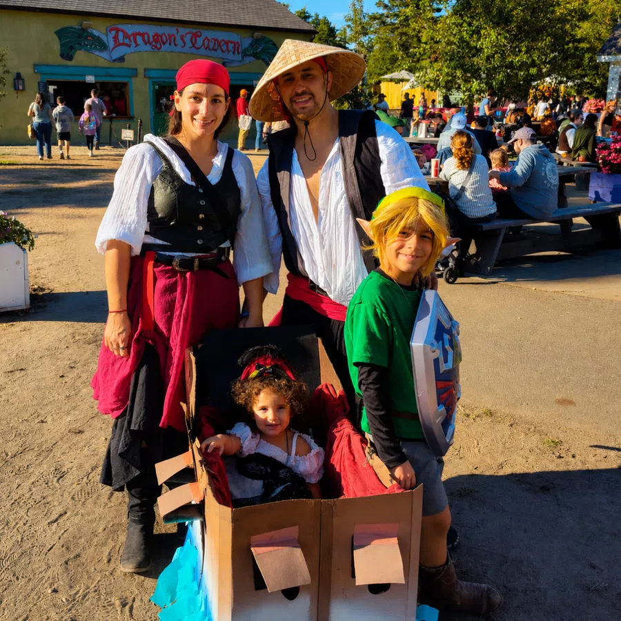 Family dressed in Renaissance and fantasy costumes posing outside Dragon's Tavern during a sunny festival day.