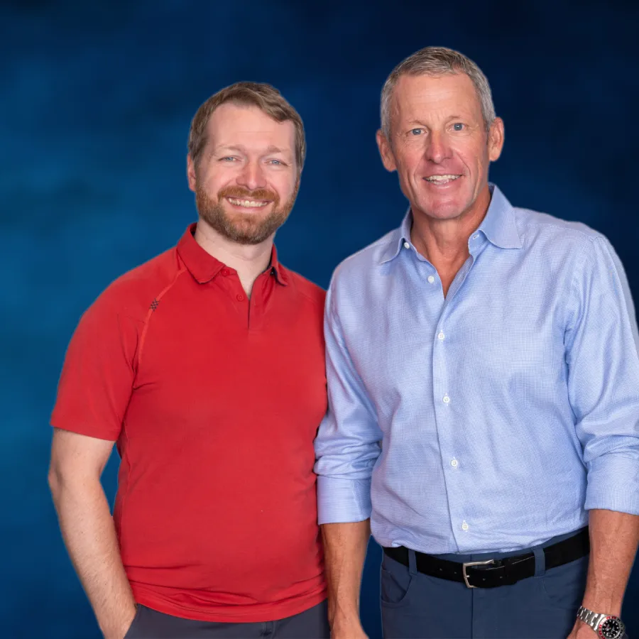 Two men standing side by side smiling against a blue gradient background, one in red polo, the other in light blue shirt.