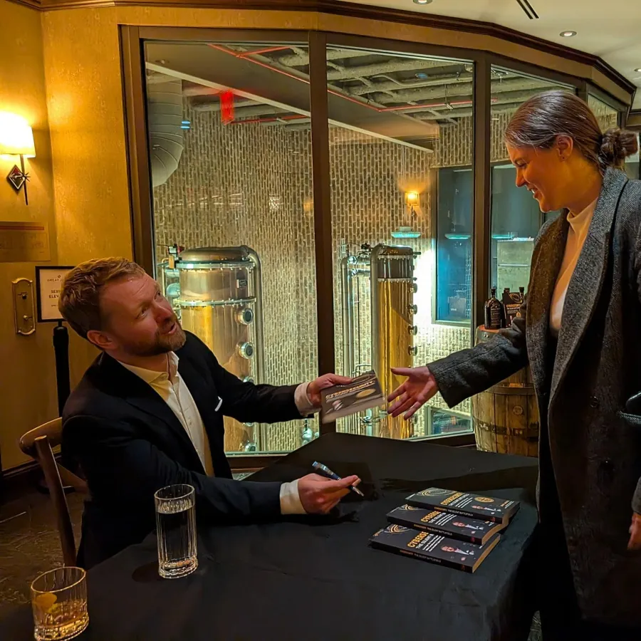Author signing books for a smiling woman at an indoor event with brewing equipment in the background.