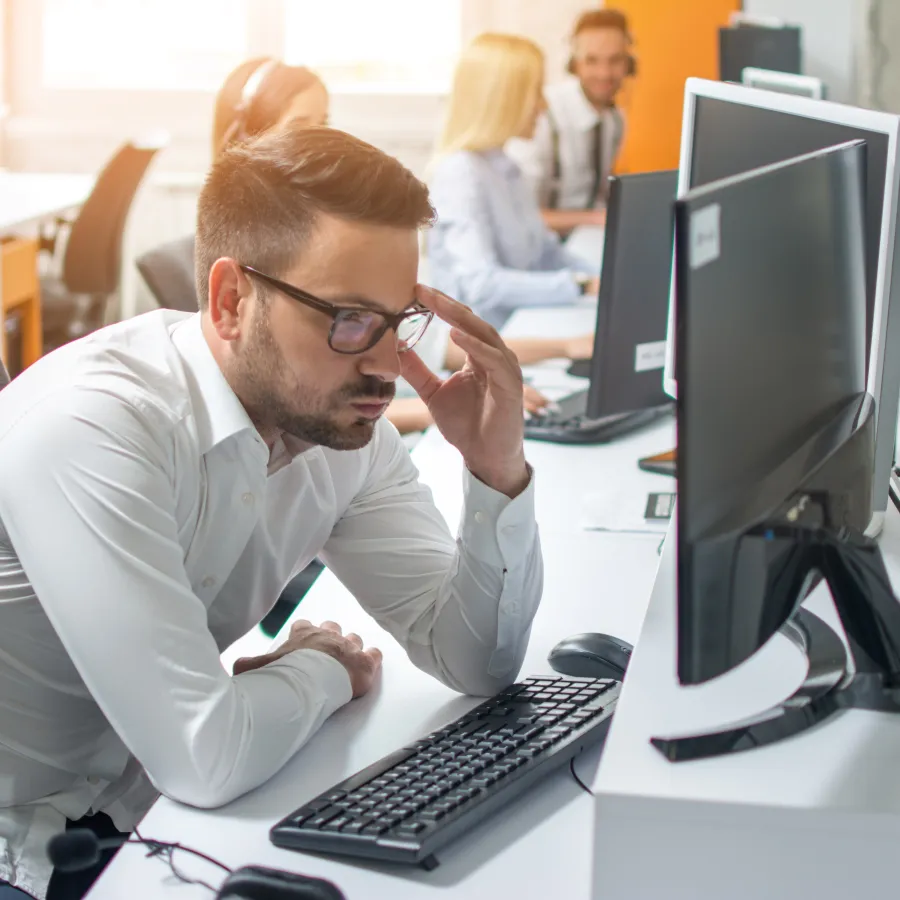 Frustrated young professional working at a computer in an office environment with colleagues in the background.