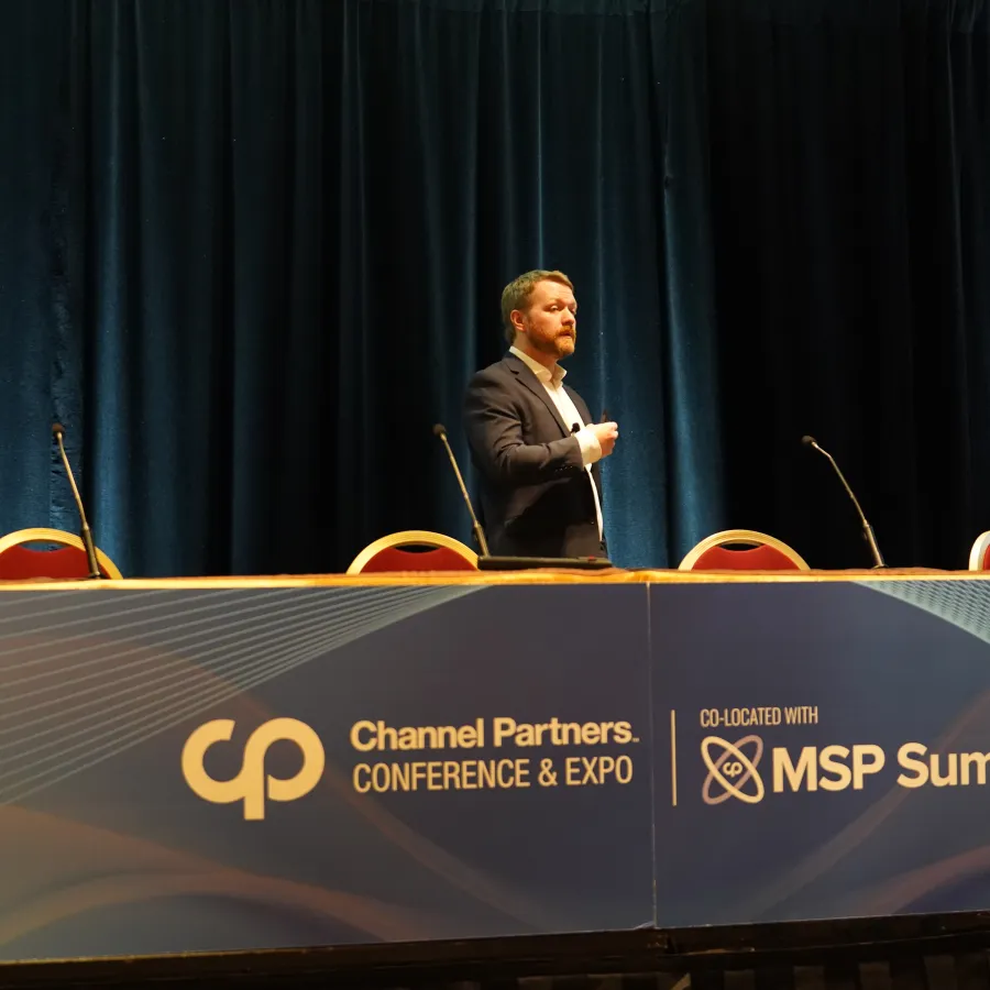 Man standing behind table at Channel Partners Conference & Expo co-located with MSP Summit with microphones and chairs.