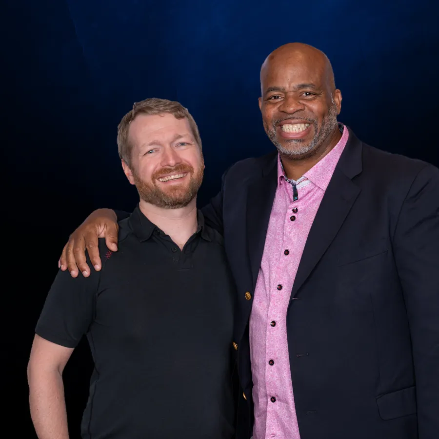 Two men smiling and posing together against a dark blue background, one in a black shirt, the other in a pink shirt with a black blazer