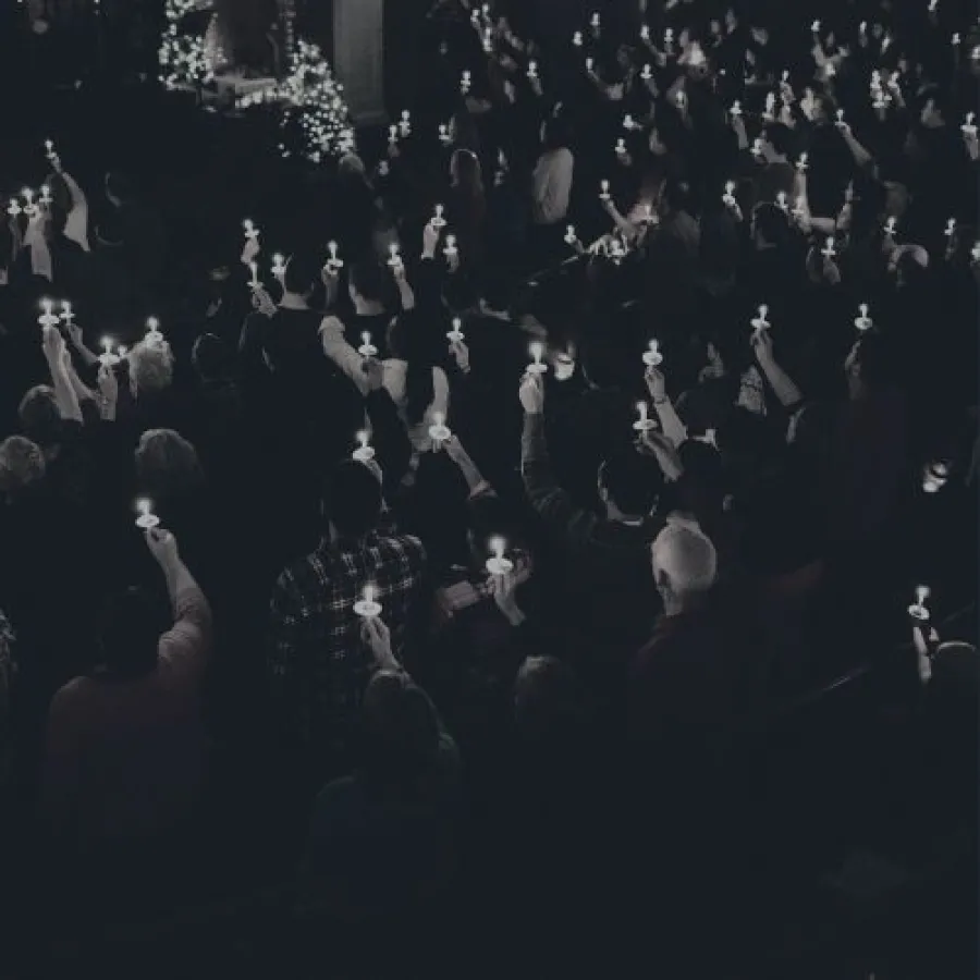 Large crowd holding lit candles in a dark setting, forming a solemn vigil or memorial gathering.