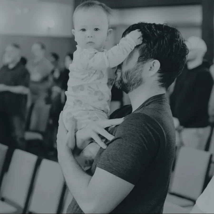 Father holding baby who touches his face in a gathering with seated and standing people in the background.