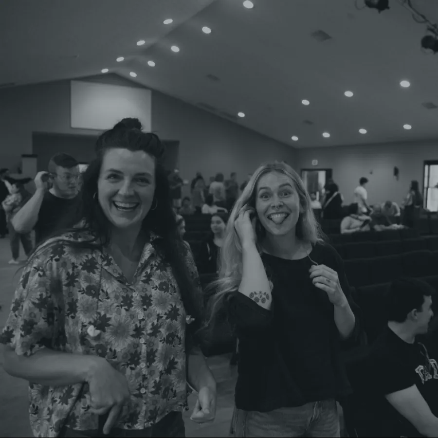 Two young women smiling and posing playfully in a crowded indoor event space with people seated and standing.
