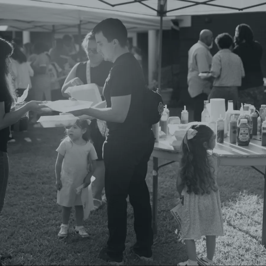 People and children gathered outdoors at a food serving table with condiments under a canopy tent.