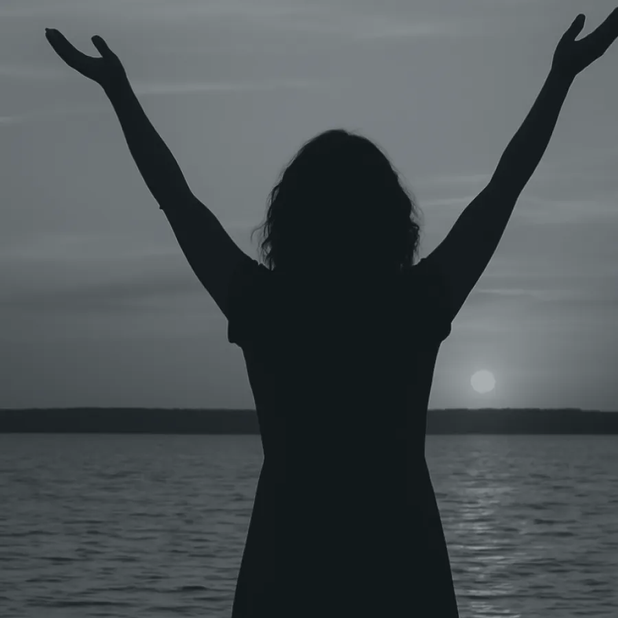 Silhouette of a person with raised arms facing a sunset over calm water and distant horizon.