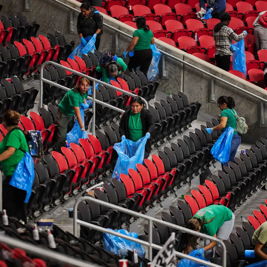 Group of people in green shirts cleaning red and black stadium seats holding blue trash bags and gloves.
