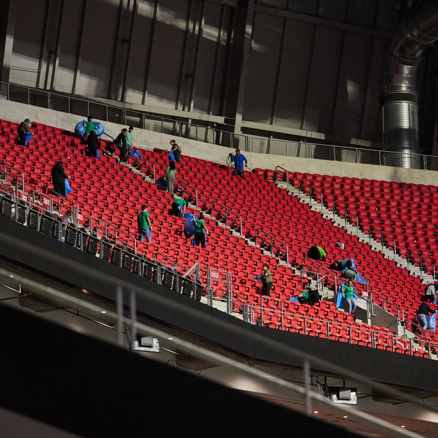 Workers in green and blue uniforms clean red stadium seats in a large indoor sports arena under bright lights.
