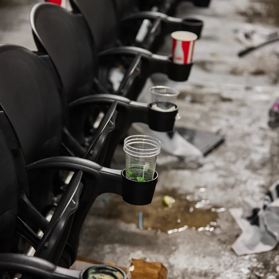 Row of black stadium seats with various empty drink cups and trash scattered on wet concrete floor.
