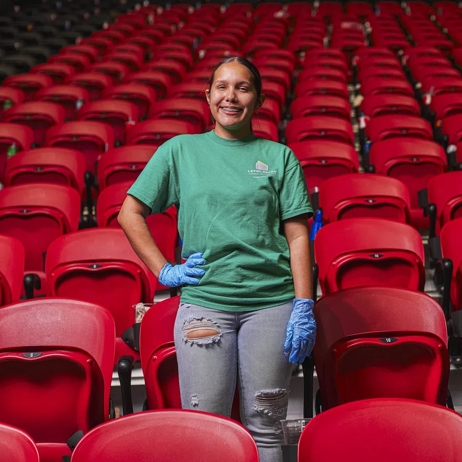 Young woman in green shirt and blue gloves standing amid rows of empty red stadium seats, smiling at camera.