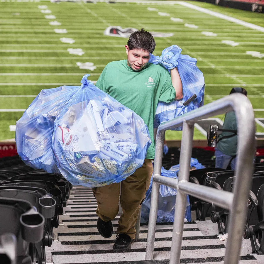 Young volunteer carries two large blue bags of recyclables up stadium stairs near a football field.