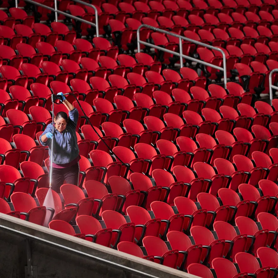 Two workers cleaning rows of empty red seats in a large indoor arena or stadium with spray bottles and cloths.