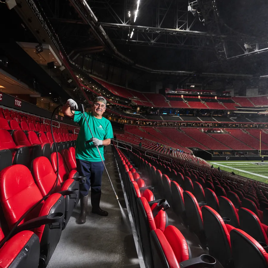 Worker sprays disinfectant on red stadium seats during cleaning inside an empty football stadium.