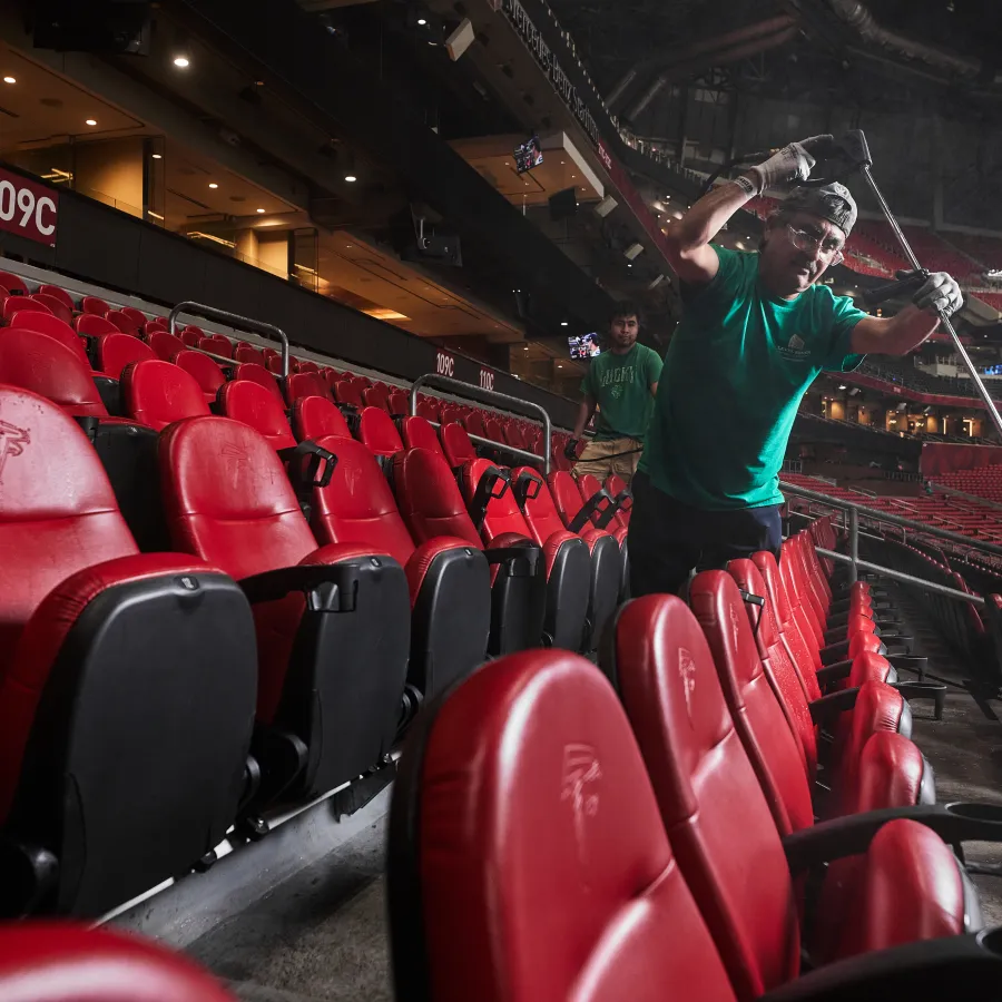 Workers cleaning and maintaining red stadium seats in an empty indoor sports arena.