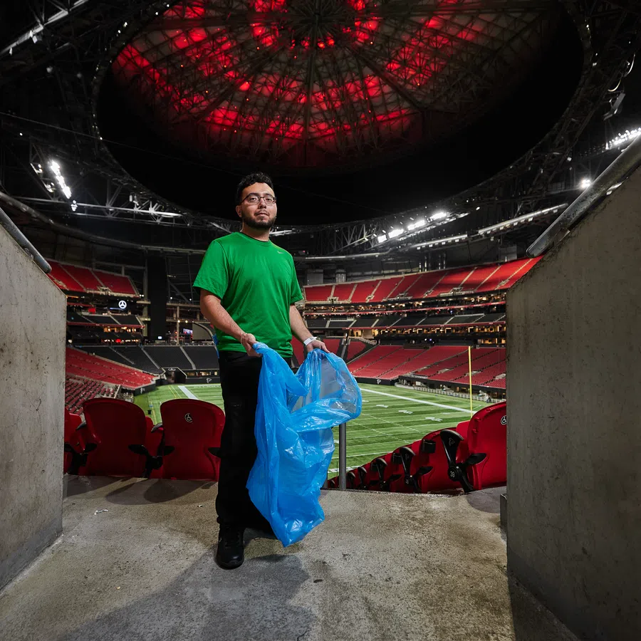 Stadium worker in green shirt holding blue trash bag inside a large football stadium with red seats and closed roof.