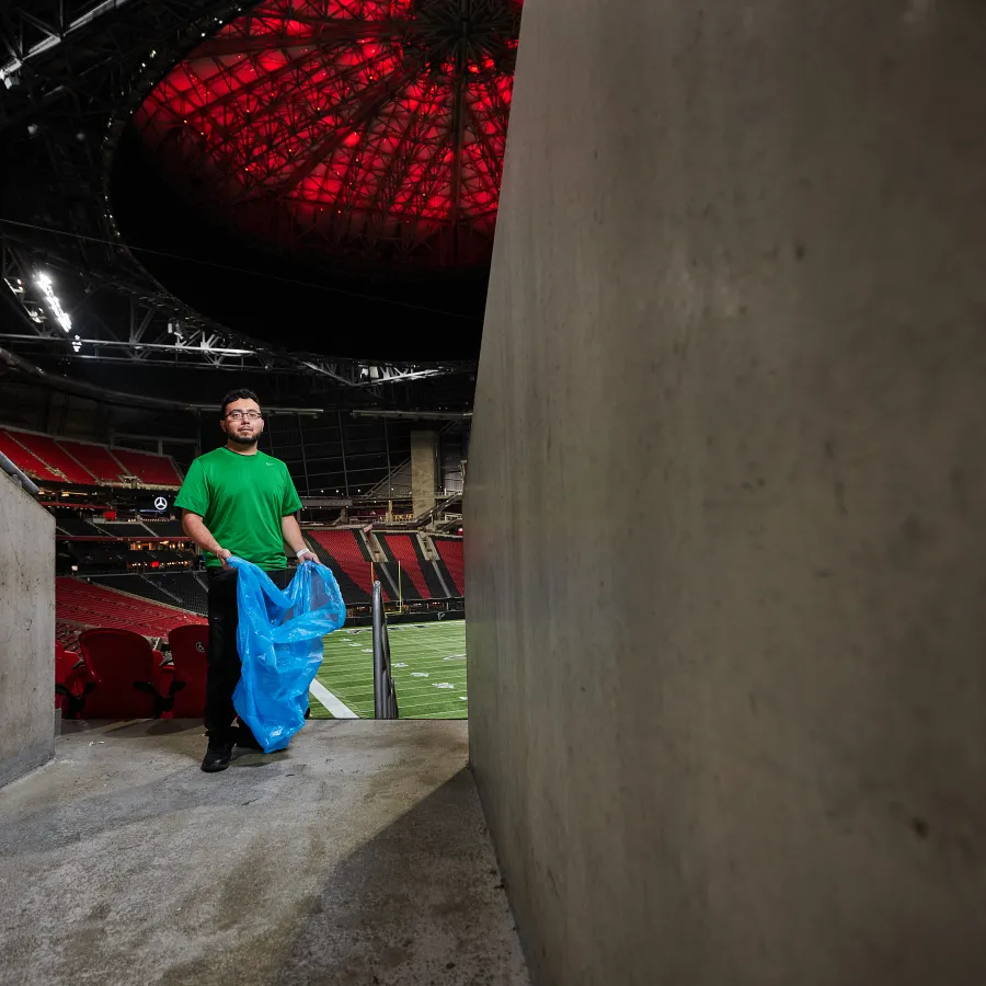 Man in green shirt holds blue trash bag in stadium tunnel with red-lit ceiling and empty seats.