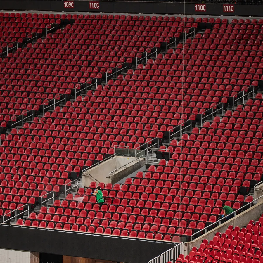 Empty red stadium seats with a few people scattered and stairways dividing sections under daylight.