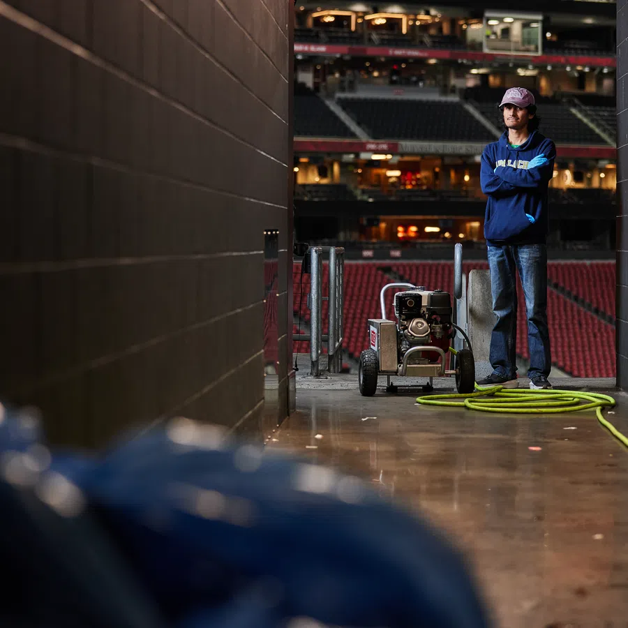 Man in blue hoodie standing in a stadium corridor next to a pressure washer and yellow hose on concrete floor.