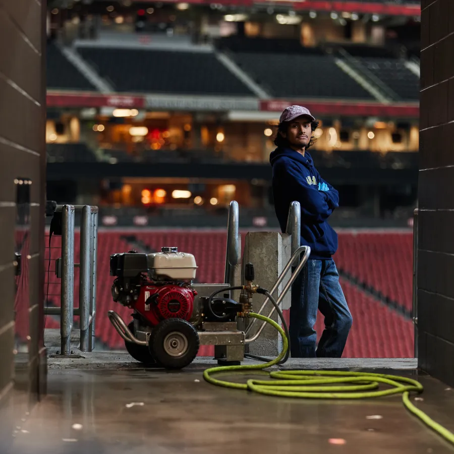 Man in casual clothes stands near equipment and coiled hose in empty stadium corridor.