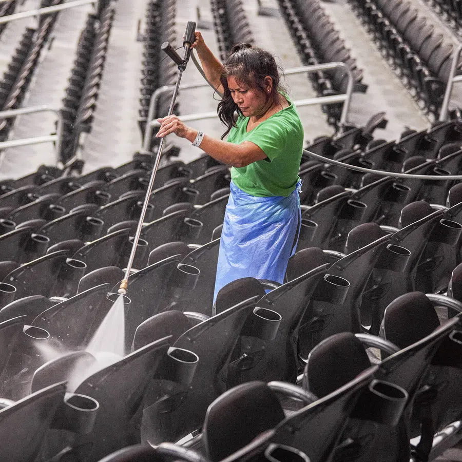 Woman in green shirt pressure washing black stadium seats in empty arena.