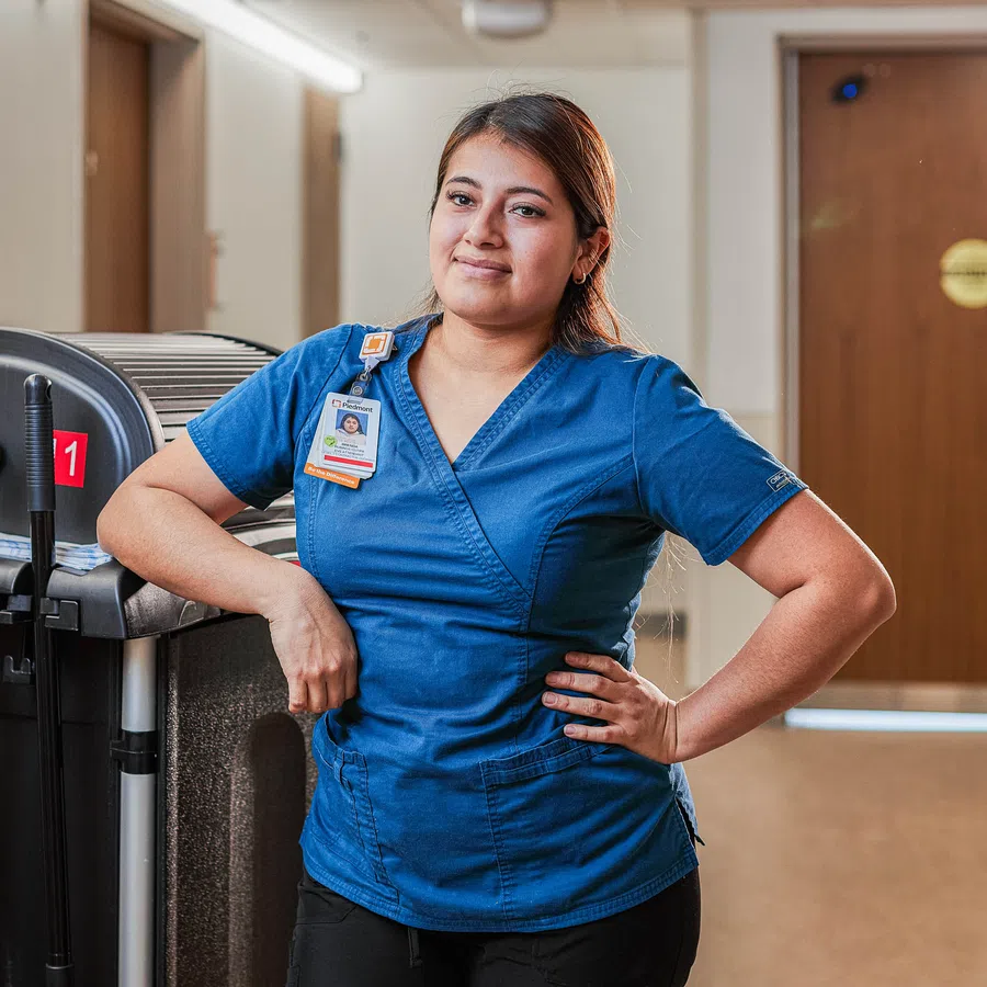 Female healthcare worker in blue scrubs standing in hospital hallway next to medical equipment cart