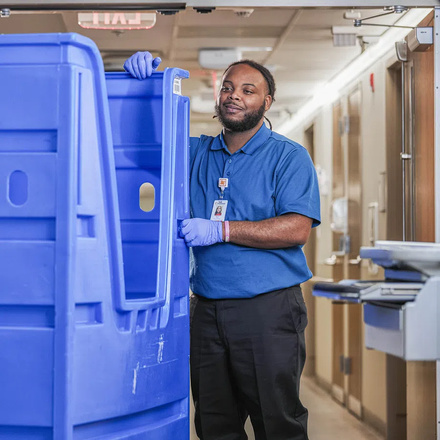 Worker in blue gloves handling large blue plastic bin in a hallway, wearing a blue shirt and ID badge.