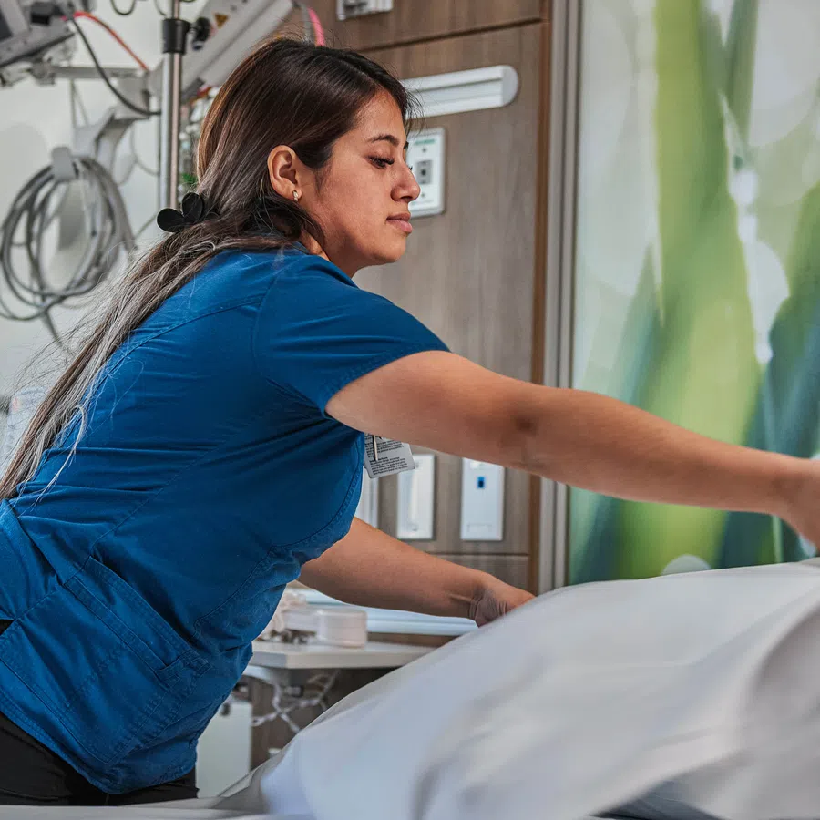 Healthcare worker in blue scrubs making a hospital bed with white linens in a patient room.
