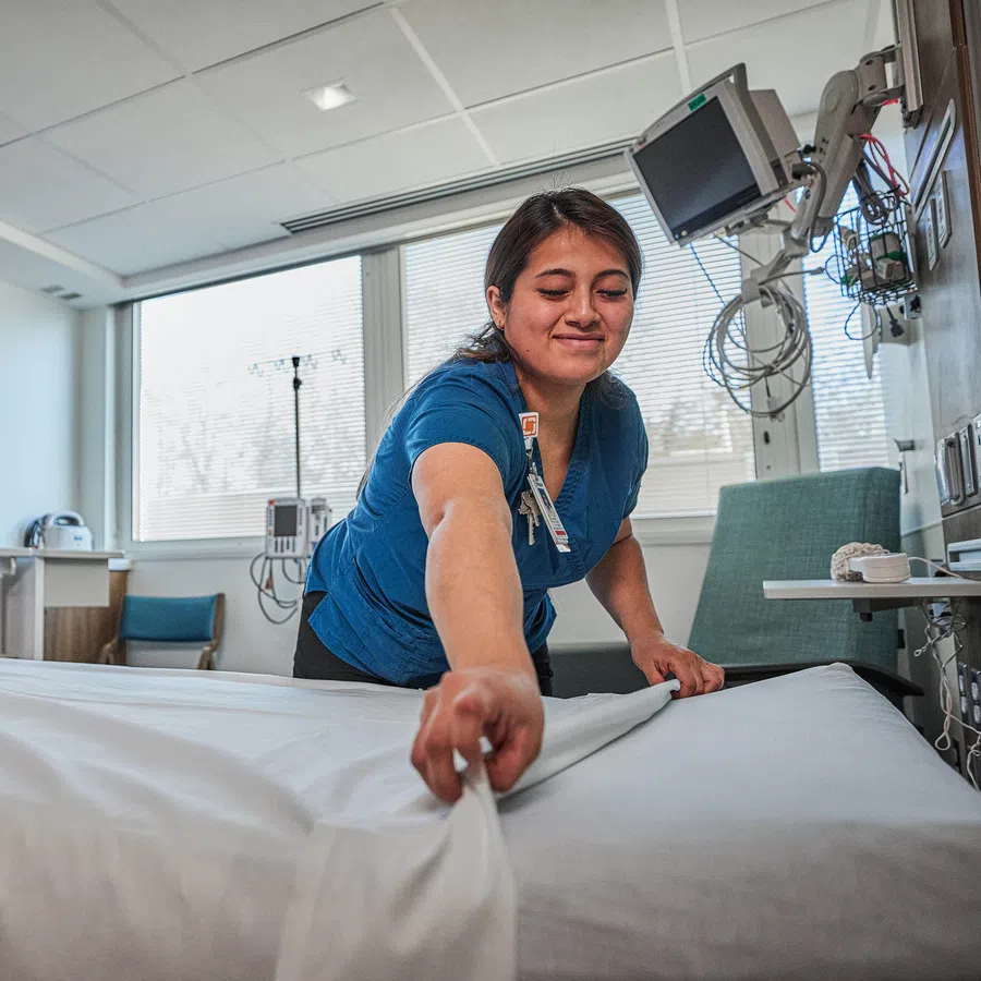 Nurse making a hospital bed in a bright patient room with medical equipment and Piedmont logo on screen.