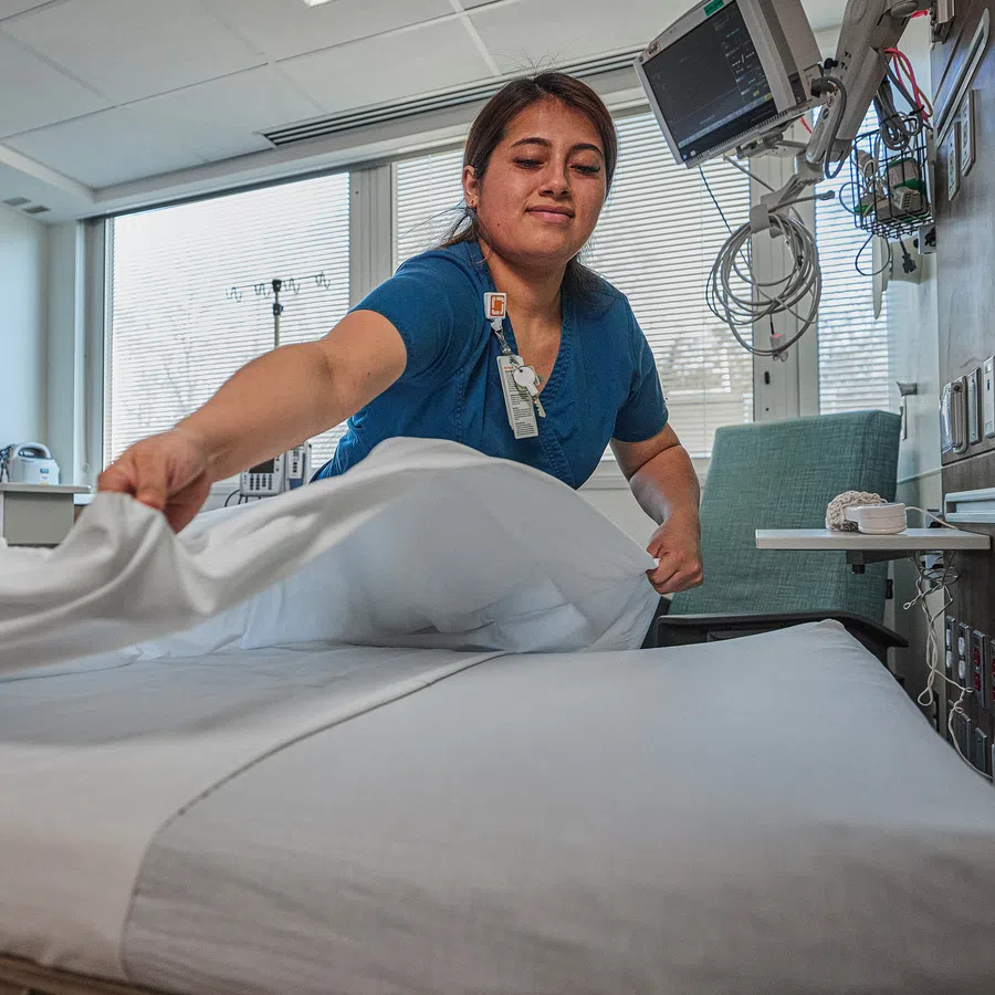 Healthcare worker changing white sheets on hospital bed in a modern patient room with medical equipment.