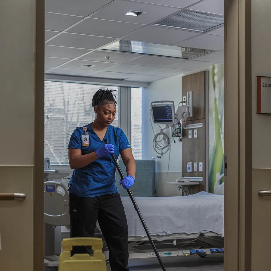 Healthcare worker in blue scrubs mopping hospital room floor with wet floor caution sign in foreground.