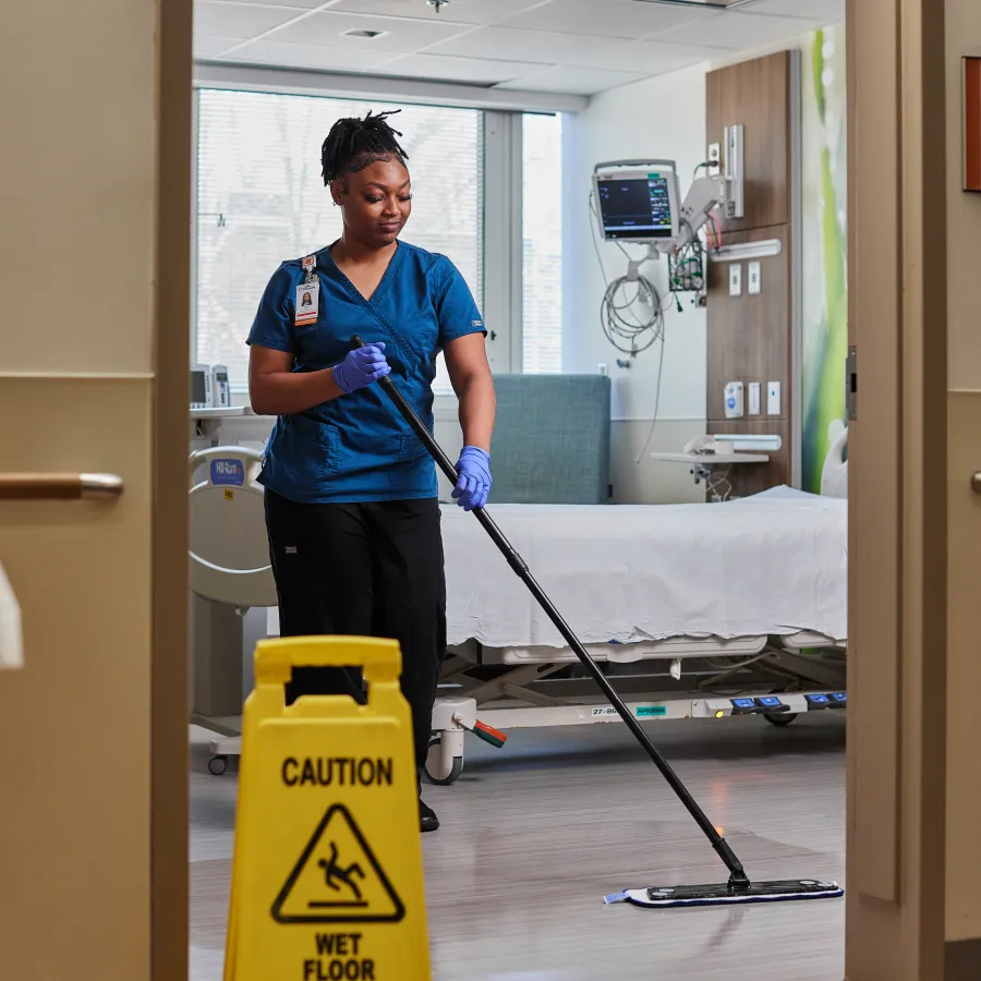 Healthcare worker mopping hospital room floor with wet floor caution sign in foreground.