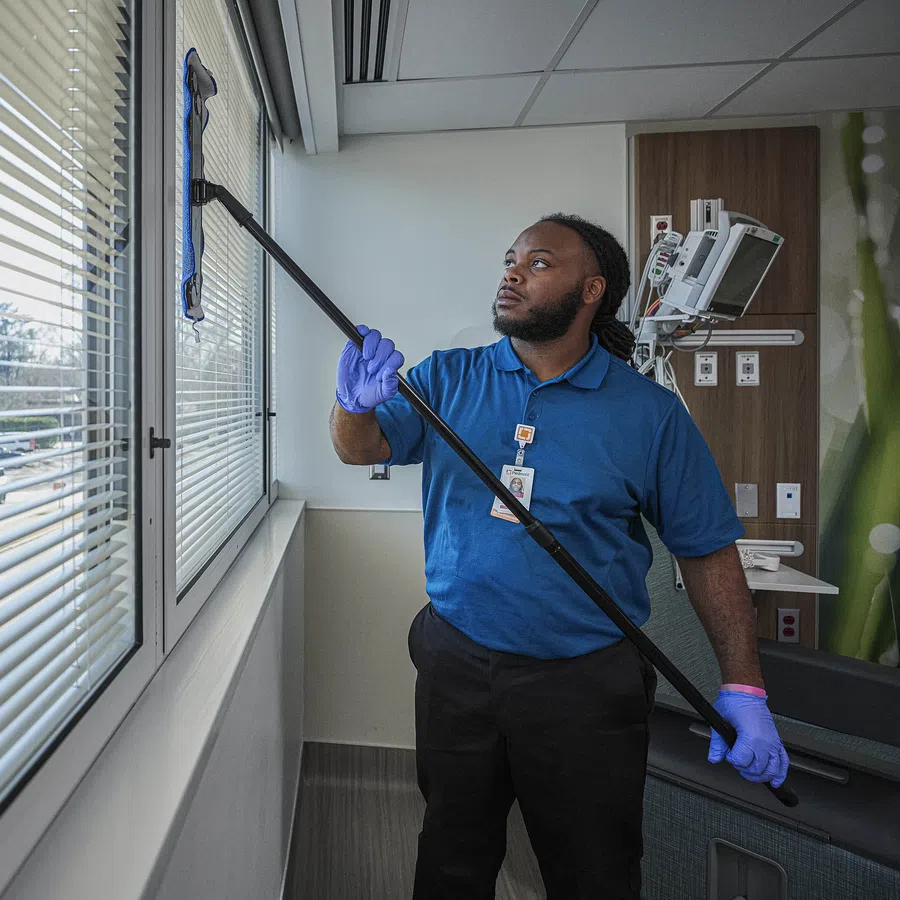 Professional cleaner wearing gloves cleaning large hospital window with mop in modern medical facility.