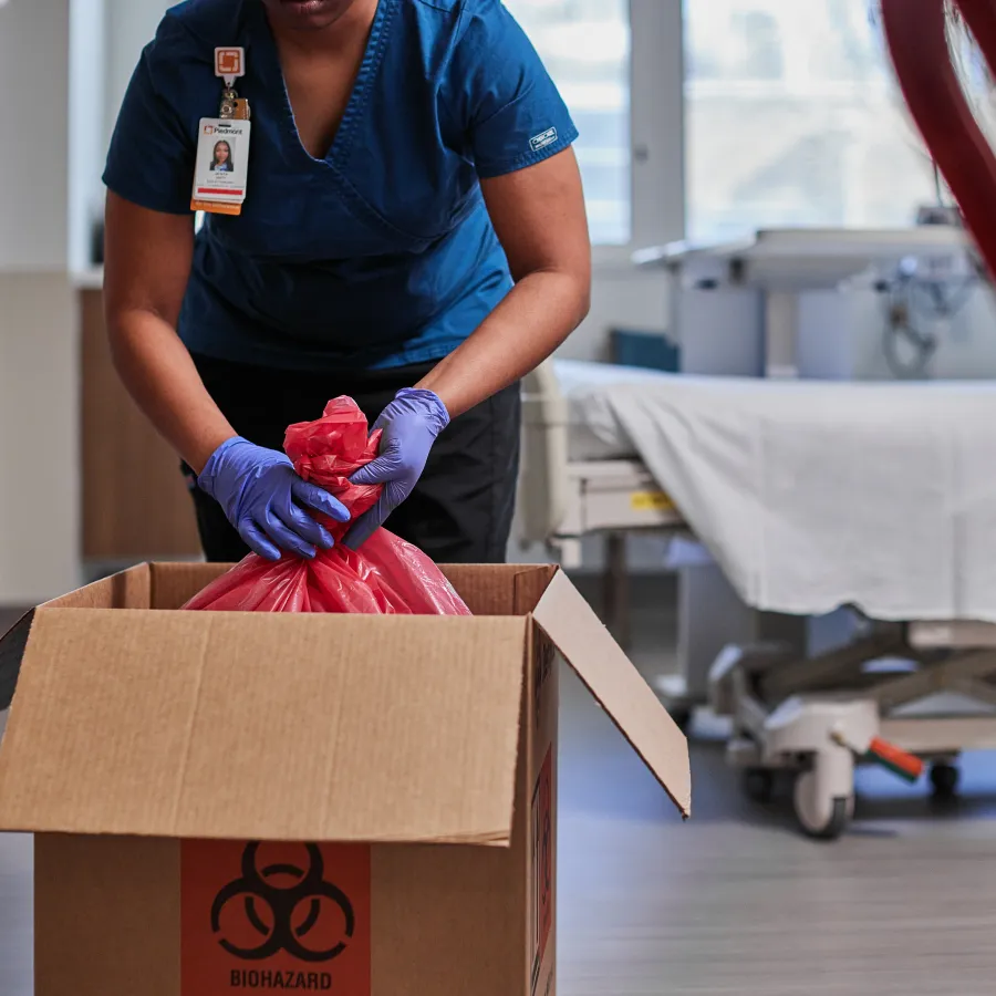 Healthcare worker in blue scrubs placing a red biohazard bag into a labeled cardboard box in a medical facility.