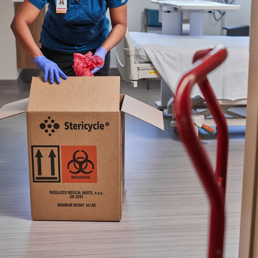 Healthcare worker in gloves disposing red biohazard bags into a Stericycle medical waste box in hospital room.