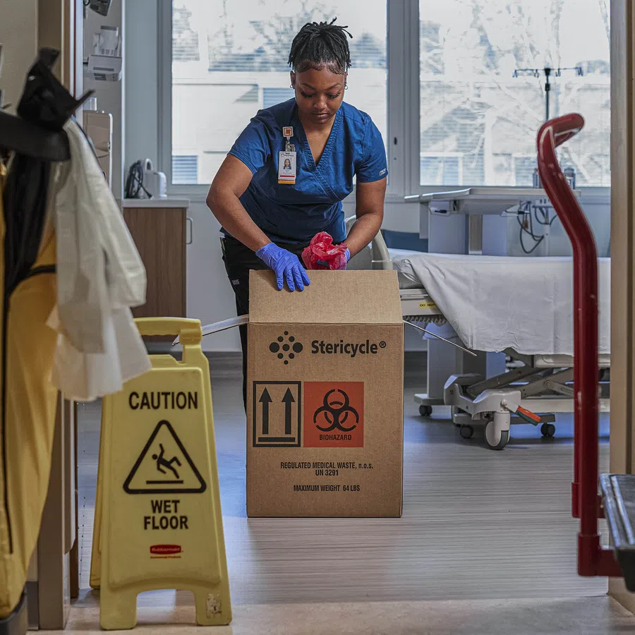 Healthcare worker wearing gloves disposing medical waste into a Stericycle biohazard box in hospital room.