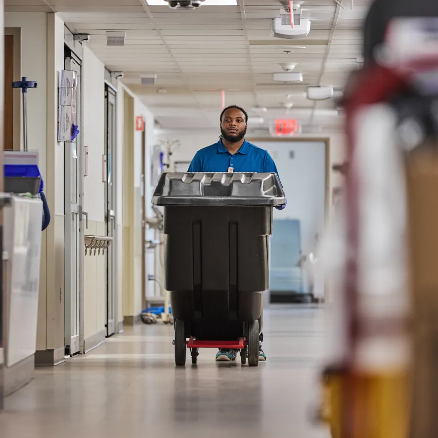 Janitor in blue uniform pushing a large black trash bin down a hospital corridor with medical equipment.