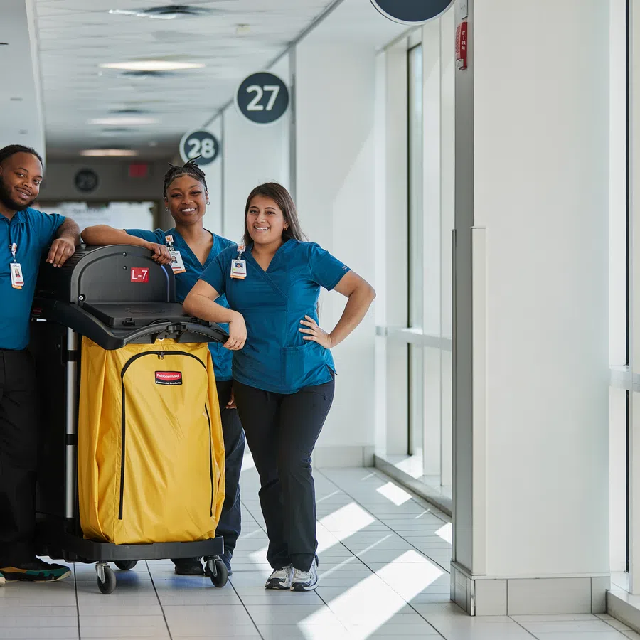 Three cleaning staff in blue uniforms standing in hallway with large yellow cleaning cart beside them.