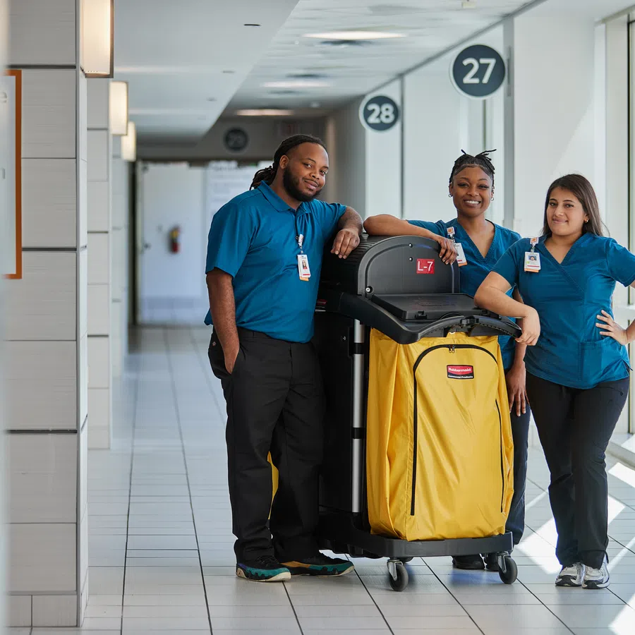 Three janitors in blue uniforms standing with a yellow cleaning cart in a bright hallway with numbered signs.