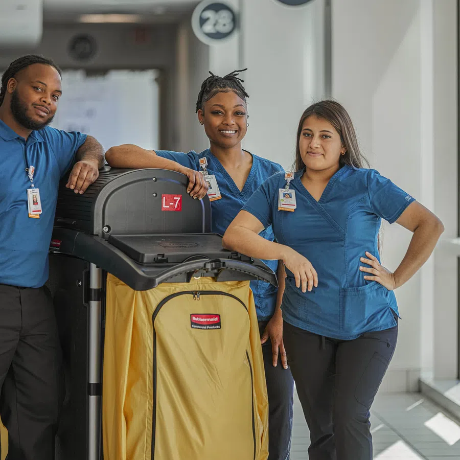 Three janitorial staff in blue uniforms pose with a yellow cleaning cart in a bright hallway.