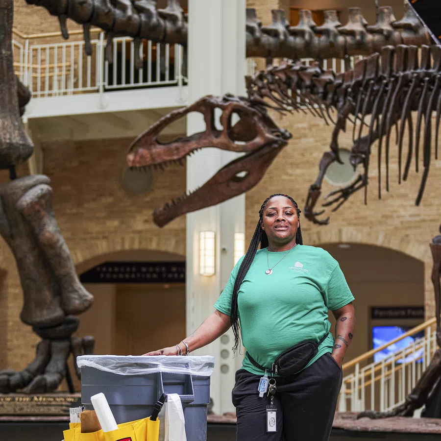 Museum janitor in green shirt standing with cleaning cart in front of dinosaur skeleton exhibit indoors.