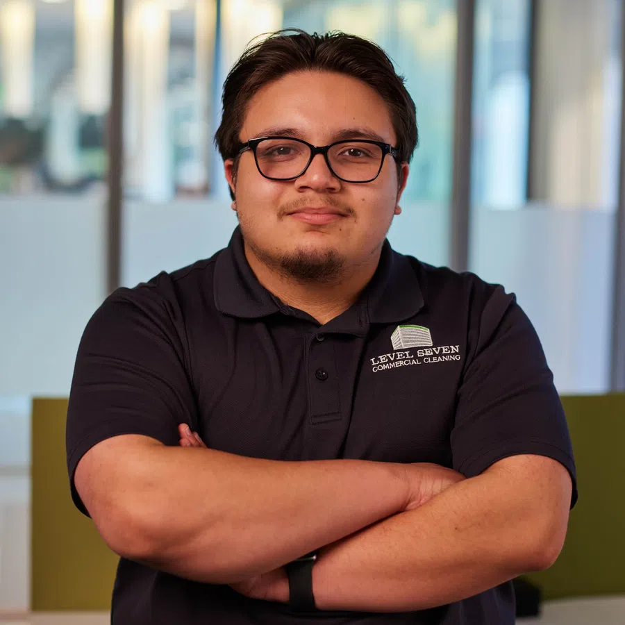 Man with glasses and black polo standing with arms crossed in modern office setting with blurred background.