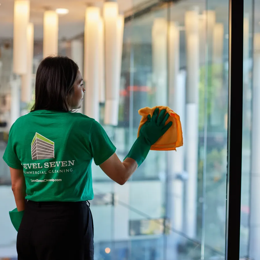 Woman wearing green Level Seven shirt cleaning glass window with a cloth inside a modern building.