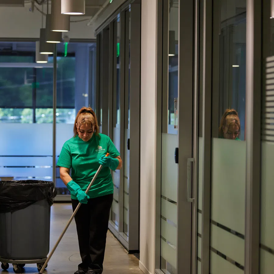 Janitor wearing green cleaning a hallway floor with mop and gloves beside trash bin in modern office space.