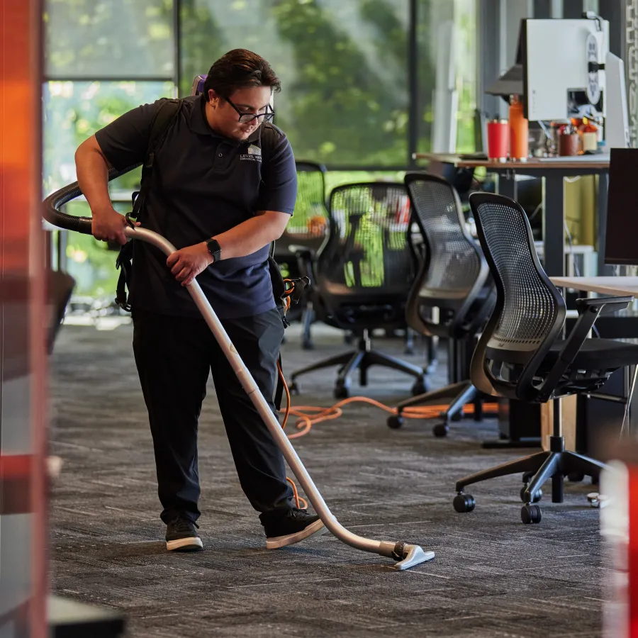 Office worker in black uniform vacuuming carpeted floor in modern workspace with chairs and desks.