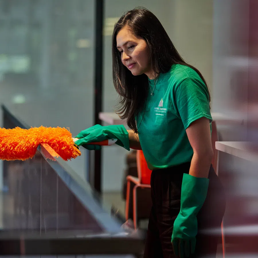 Woman in green shirt and gloves dusting a glass surface with an orange duster in an indoor setting