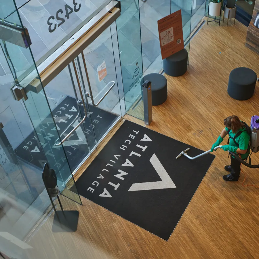 Person vacuuming wooden floor near glass entrance with Atlanta Tech Village logo mat inside building lobby
