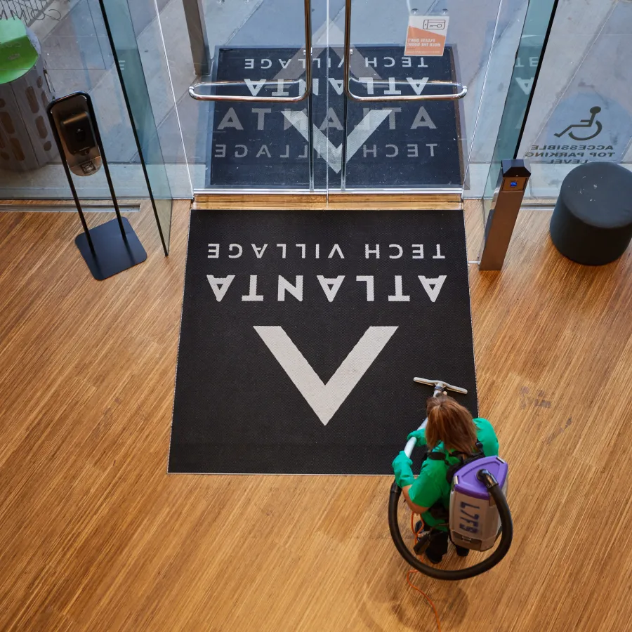 Woman cleaning hardwood floor near glass doors with Atlanta Tech Village logo mat in front entrance.
