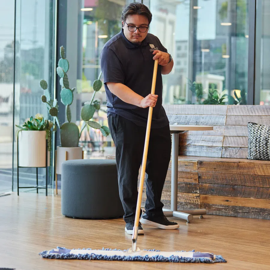 Man mopping wooden floor in bright modern cafe with indoor plants and large glass windows during daytime.